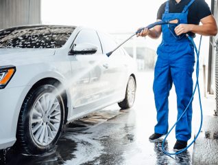 Professional washer in blue uniform washing luxury car with water gun on an open air car wash. Close up photo.