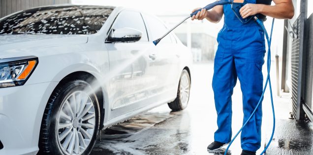 Professional washer in blue uniform washing luxury car with water gun on an open air car wash. Close up photo.